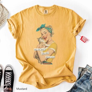 Puede incluir: Camiseta de color mostaza con un gr&aacute;fico retro de una mujer sosteniendo un gato, con el texto "my cat and I talk shit about you". La camiseta tiene mangas cortas y cuello redondo. Una etiqueta que dice "Guts Tees" est&aacute; adjunta.