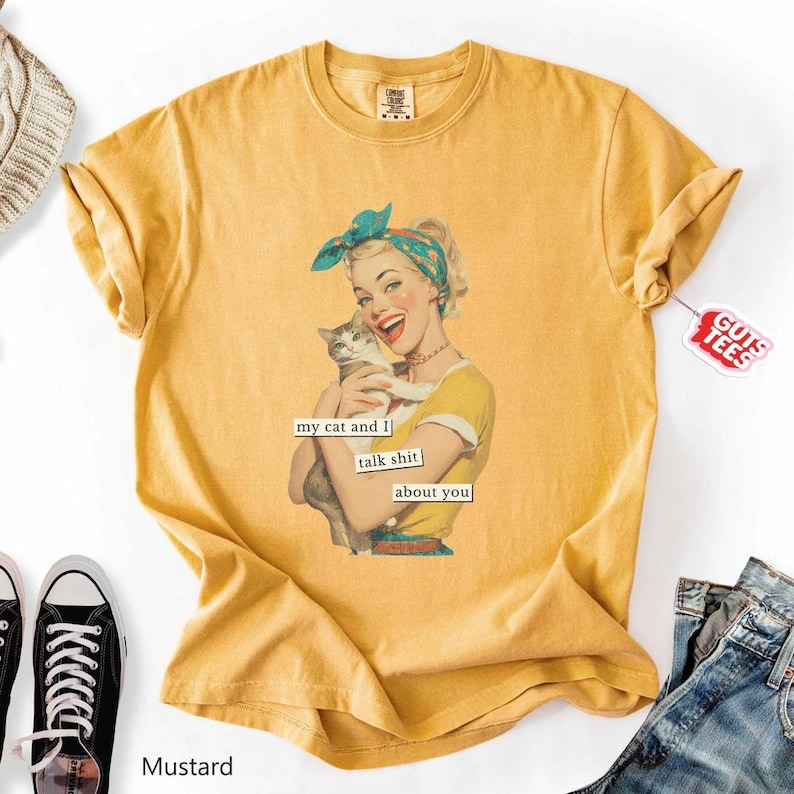 Puede incluir: Camiseta de color mostaza con un gr&aacute;fico retro de una mujer sosteniendo un gato, con el texto "my cat and I talk shit about you". La camiseta tiene mangas cortas y cuello redondo. Una etiqueta que dice "Guts Tees" est&aacute; adjunta.