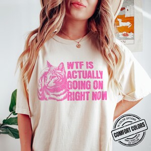 May include: A white t-shirt with a pink graphic of a cat and the text "WTF IS ACTUALLY GOING ON RIGHT NOW". The shirt has a pink label that says "COMFORT COLORS" with the text "100% RING-SPUN US COTTON" underneath.
