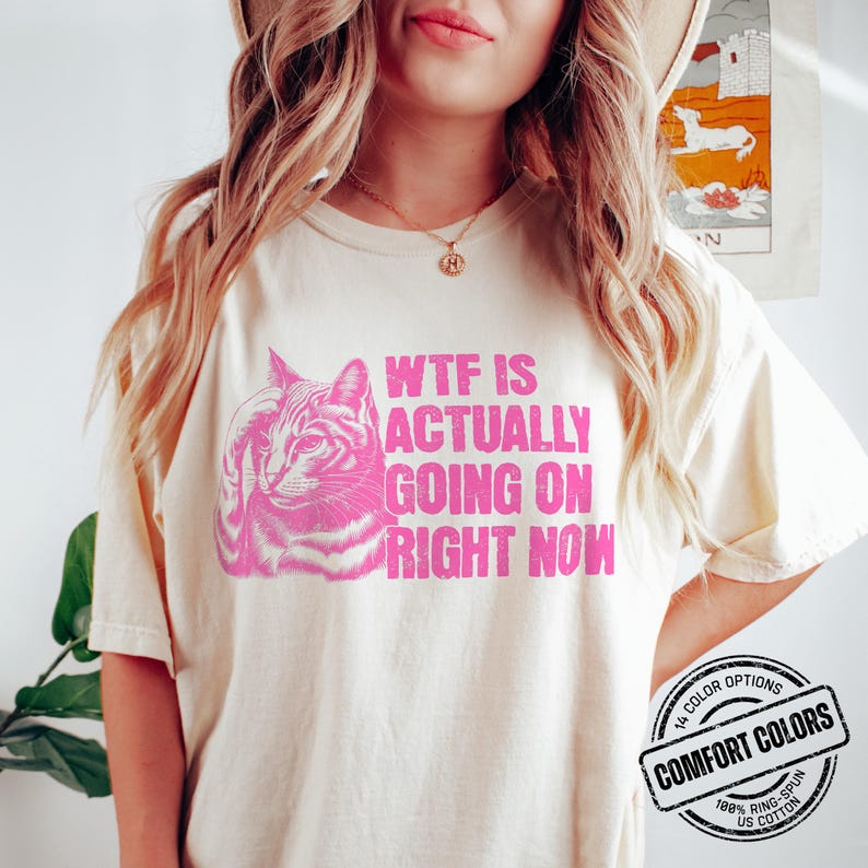 May include: A white t-shirt with a pink graphic of a cat and the text "WTF IS ACTUALLY GOING ON RIGHT NOW". The shirt has a pink label that says "COMFORT COLORS" with the text "100% RING-SPUN US COTTON" underneath.