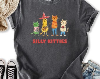 May include: Dark gray t-shirt featuring a whimsical illustration of four cartoon cats. The cats are in various poses, with one playing a guitar. The text "SILLY KITTIES" is printed in red below the cats.