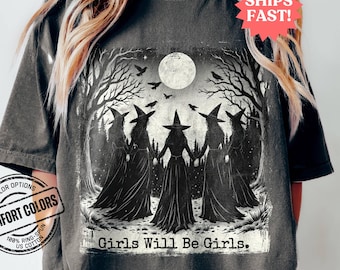 May include: A dark grey t-shirt with a graphic of four witches standing in a circle under a full moon in a forest. The text "Girls Will Be Girls." is printed below the graphic.