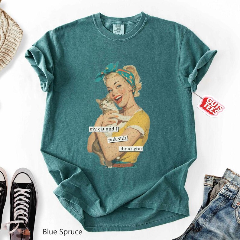Puede incluir: Camiseta azul abeto con una ilustraci&oacute;n retro de una mujer sosteniendo un gato, con el texto "my cat and I talk shit about you". La camiseta tiene un corte relajado y una etiqueta "Guts Tees".