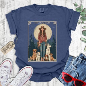 May include: A navy blue t-shirt with the text "The Dog Mom" in white lettering. The design features a woman with long brown hair and a white dog, surrounded by other dogs in a woodland setting. The moon is visible in the background.