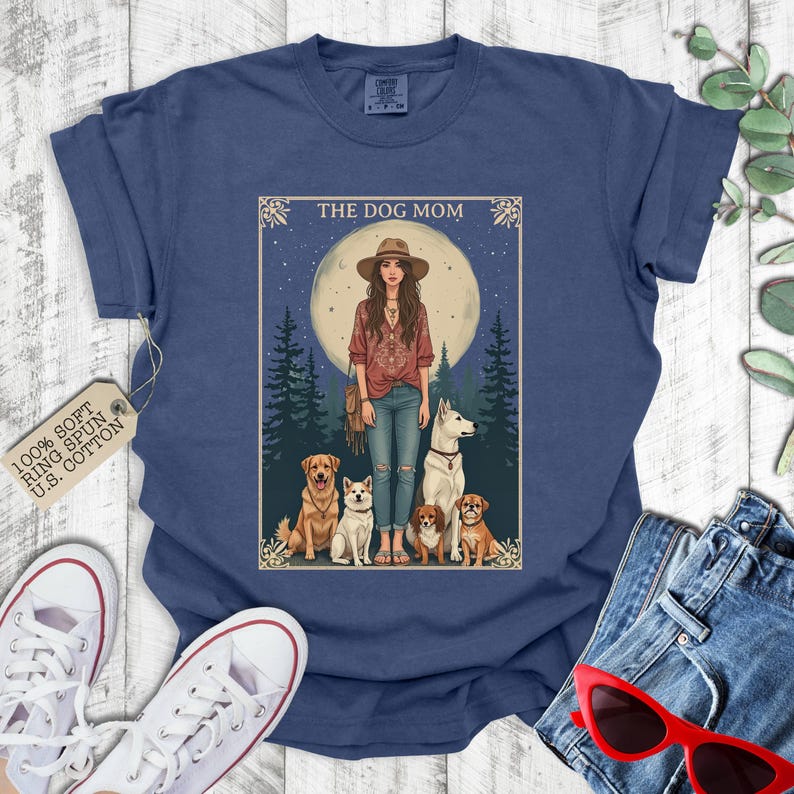 May include: A navy blue t-shirt with the text "The Dog Mom" in white lettering. The design features a woman with long brown hair and a white dog, surrounded by other dogs in a woodland setting. The moon is visible in the background.