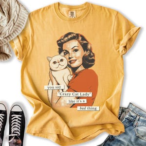May include: Mustard yellow t-shirt featuring a vintage-style illustration of a woman holding a white cat. The text reads "you say 'Crazy Cat Lady' like it's a bad thing". The shirt has a crew neck and short sleeves.