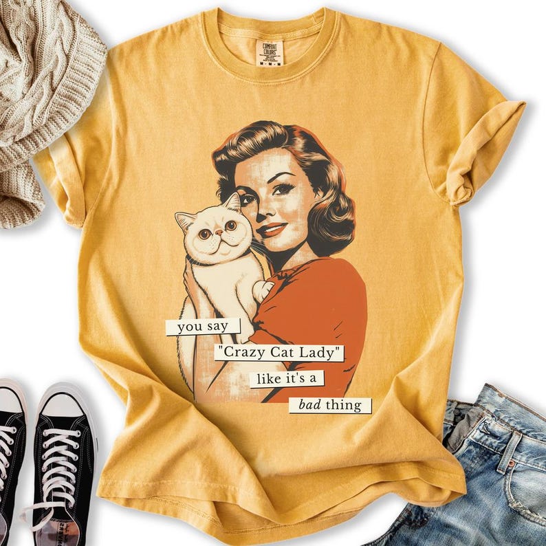 May include: Mustard yellow t-shirt featuring a vintage-style illustration of a woman holding a white cat. The text reads "you say 'Crazy Cat Lady' like it's a bad thing". The shirt has a crew neck and short sleeves.