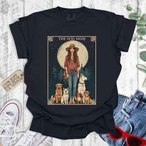 May include: A black t-shirt featuring a vintage-style illustration of a woman with long brown hair and four dogs. The text "The Dog Mom" is at the top of the illustration.