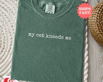 May include: A green t-shirt with the phrase "my cat kneads me" in white lettering. The shirt has a crew neck and a tag that says "Comfort Colors". The shirt is folded on a white rug.