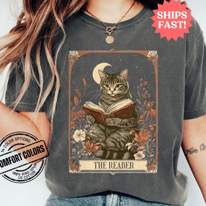 May include: A vintage-style tarot card illustration featuring a grey tabby cat reading a book. The card is surrounded by flowers and has the text "THE READER" at the bottom.