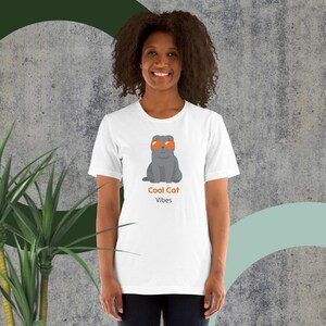 Puede incluir: Camiseta blanca con un gato gris de dibujos animados con gafas de sol naranjas. El texto "Cool Cat Vibes" está impreso debajo del gato. Una prenda informal para el día a día.