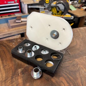 May include: A clear, rectangular router base plate with a metal insert and a set of metal bushings in a black foam insert. The items are for woodworking and are displayed on a wooden surface.