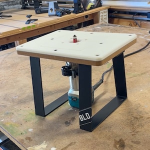 Puede incluir: Una mesa de enrutador blanca con patas de metal negro. La mesa de enrutador est&aacute; sobre una superficie de madera. La mesa de enrutador tiene una fresa instalada y un enrutador est&aacute; conectado a la mesa.