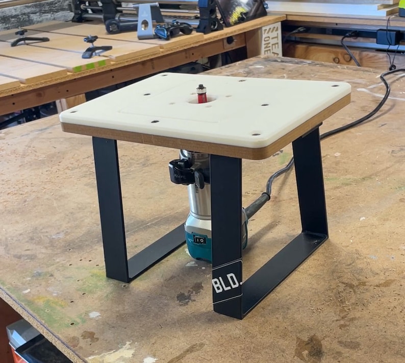 Puede incluir: Una mesa de enrutador blanca con patas de metal negro. La mesa de enrutador est&aacute; sobre una superficie de madera. La mesa de enrutador tiene una fresa instalada y un enrutador est&aacute; conectado a la mesa.