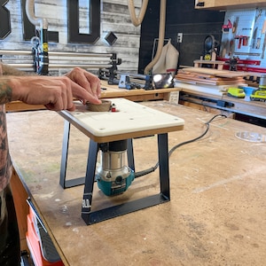 Puede incluir: Una mesa de enrutador blanca con una base de metal negro y un enrutador turquesa adjunto. La mesa est&aacute; sobre un banco de trabajo de madera en un taller.