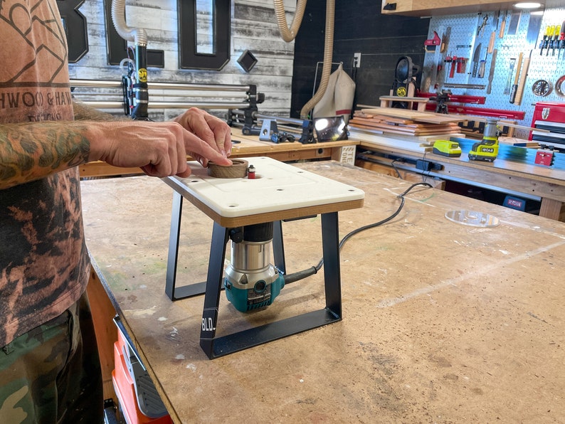 Puede incluir: Una mesa de enrutador blanca con una base de metal negro y un enrutador turquesa adjunto. La mesa est&aacute; sobre un banco de trabajo de madera en un taller.