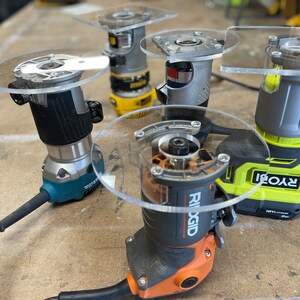 Puede incluir: Cuatro marcas diferentes de fresadoras el&eacute;ctricas con bases de pl&aacute;stico transparente. Las fresadoras est&aacute;n sobre una superficie de madera. Las marcas son Ryobi, Ridgid, DeWalt y otra marca que no es visible.