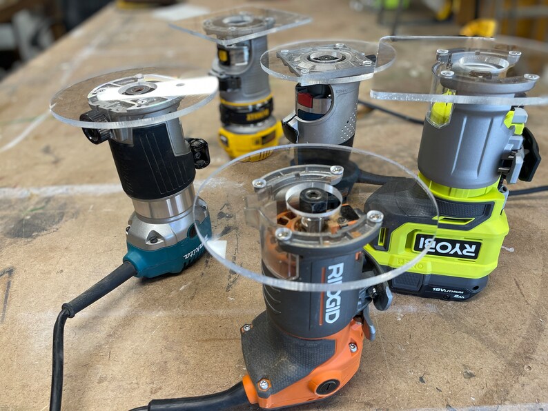 Puede incluir: Cuatro marcas diferentes de fresadoras el&eacute;ctricas con bases de pl&aacute;stico transparente. Las fresadoras est&aacute;n sobre una superficie de madera. Las marcas son Ryobi, Ridgid, DeWalt y otra marca que no es visible.