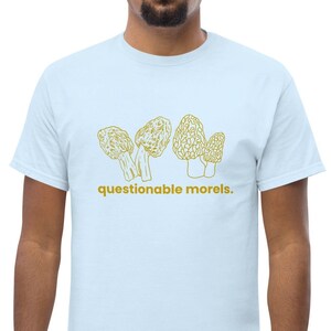 May include: A light blue t-shirt featuring a graphic of three yellow-gold outlined morel mushrooms and the text "questionable morels."