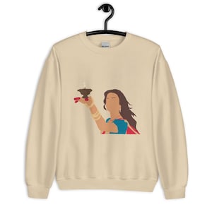 May include: Beige sweatshirt featuring a graphic of a person holding a lit lamp. The person is wearing a blue and red outfit with gold jewelry. The design is a minimalist illustration style.