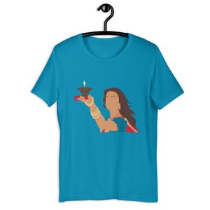 May include: A turquoise t-shirt featuring a graphic of a person holding a lit oil lamp. The person is wearing a red and blue sari, gold bangles, and a necklace. The design is a minimalist illustration.