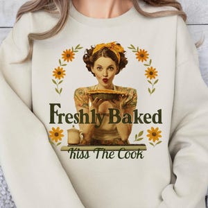 May include: Cream sweatshirt with a retro graphic of a woman holding a pie, encircled by sunflowers. The text reads "Freshly Baked" and "Kiss The Cook". The woman in the graphic wears a yellow headscarf and apron, in a vintage style.