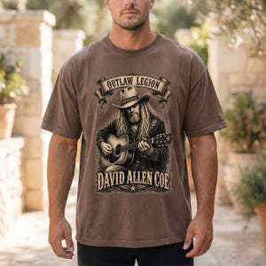 May include: Brown t-shirt with a graphic of a man playing a guitar, the text reads "OUTLAW LEGION" and "DAVID ALLEN COE". The design is in a vintage style.