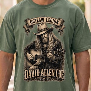 May include: Sage green t-shirt with a detailed graphic of a man with long hair and a cowboy hat playing an acoustic guitar. The shirt includes the text "OUTLAW LEGION" and "DAVID ALLEN COE" in a vintage style.