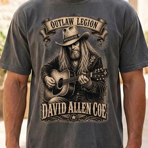 May include: Grey t-shirt with a graphic of a man playing a guitar, wearing a cowboy hat. The text reads "OUTLAW LEGION" above and "DAVID ALLEN COE" below the image, with a star emblem.
