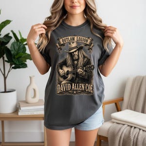 May include: Charcoal grey t-shirt with a detailed illustration of a man playing a guitar, wearing a cowboy hat. The text "OUTLAW LEGION" is above the illustration, and "DAVID ALLEN COE" is below. The shirt is a comfortable, casual style.
