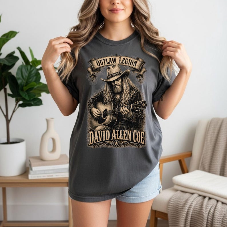 May include: Charcoal grey t-shirt with a detailed illustration of a man playing a guitar, wearing a cowboy hat. The text "OUTLAW LEGION" is above the illustration, and "DAVID ALLEN COE" is below. The shirt is a comfortable, casual style.