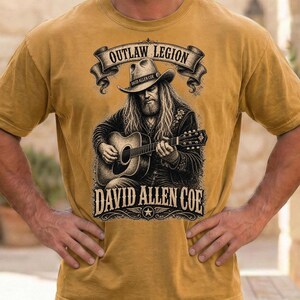 May include: Mustard yellow t-shirt with a graphic of a man playing a guitar, wearing a cowboy hat. The text "OUTLAW LEGION" is above the image, and "DAVID ALLEN COE" is below. The shirt has short sleeves.