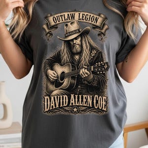 May include: Charcoal grey t-shirt with a detailed illustration of a man playing a guitar, wearing a cowboy hat. The text reads "OUTLAW LEGION" above and "DAVID ALLEN COE" below the image, with a star emblem.