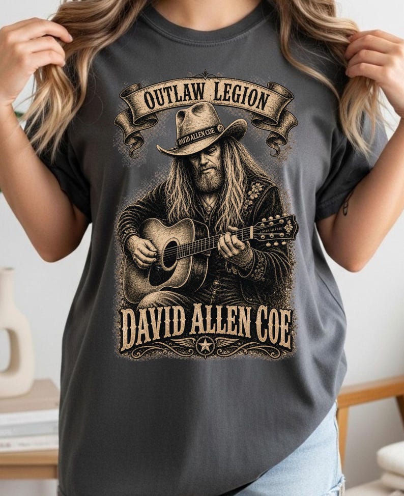 May include: Charcoal grey t-shirt with a detailed illustration of a man playing a guitar, wearing a cowboy hat. The text reads "OUTLAW LEGION" above and "DAVID ALLEN COE" below the image, with a star emblem.