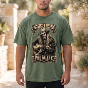 May include: Olive green t-shirt with a graphic of a musician playing a guitar, and the text "OUTLAW LEGION" above and "DAVID ALLEN COE" below. The design is in brown and black.