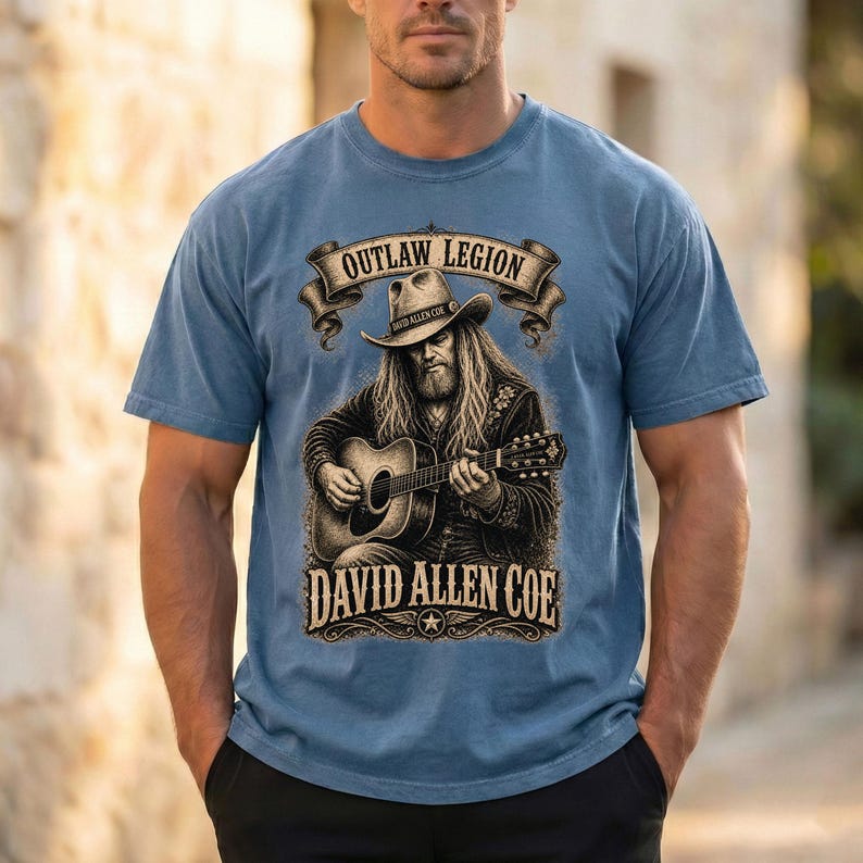 May include: Blue short-sleeved t-shirt with a graphic of a musician playing a guitar. The graphic includes the text "OUTLAW LEGION" and "DAVID ALLEN COE" in a vintage style. The musician is wearing a cowboy hat.