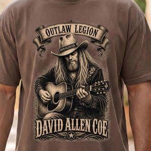 May include: Brown t-shirt with a detailed illustration of David Allen Coe playing a guitar. The design includes the text "OUTLAW LEGION" above and "DAVID ALLEN COE" below the image, with a star emblem.
