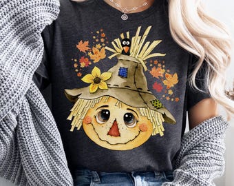 Könnte beinhalten: Dunkelgraues T-Shirt mit einem freundlichen Vogelscheuchengesicht, Strohhut, gelber Blume und Herbstblättern. Ein kleiner schwarzer Vogel sitzt auf dem Hut. Ein grafisches T-Shirt zum Thema Herbst.