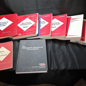 May include: A collection of technical reference books including several red-covered Structural Welding Code Steel manuals, a smaller red Welding in Building Construction book, and a black Procedure Handbook of Arc Welding by Lincoln Electric.