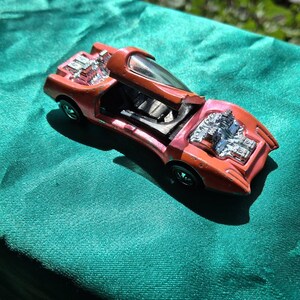 May include: A vintage orange die-cast toy car featuring a hinged canopy roof that opens upward, exposed chrome-colored engine details at the front and rear, and black wheels. The paint shows signs of wear and chipping across the body.