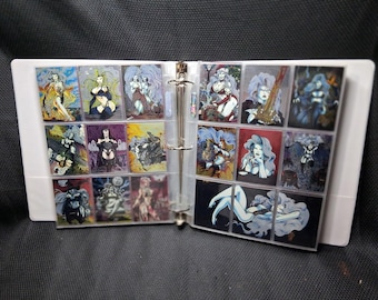 May include: A white three-ring binder containing multiple clear plastic sleeves filled with collectible trading cards. The cards feature various colorful fantasy-themed illustrations of characters with long hair, swords, and dark attire in a grid layout.