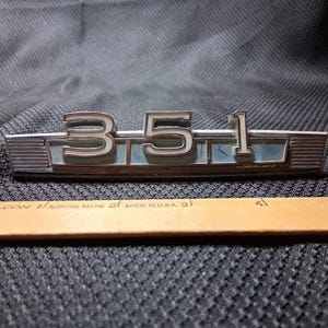 Puede incluir: Un emblema automotriz de metal plateado con los números 351 en relieve sobre un fondo rectangular azul claro con acentos laterales acanalados, colocado sobre una regla de madera que mide aproximadamente 12,7 cm de largo.