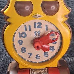 May include: A yellow owl-shaped clock with brown accents and a cream-colored clock face. The clock features blue numerals, a red clock hand, and the text "Answer Clock by TOMY". Two red buttons are located at the base.