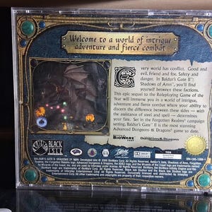 May include: The back of a Baldur's Gate II: Shadows of Amn PC game case. The case has a dark blue and gold design with the text "Welcome to a world of intrigue adventure and fierce combat". The case features a dragon illustration.