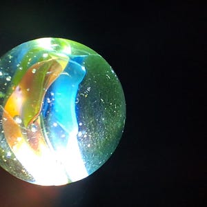 Può includere: Una biglia di vetro trasparente con vortici interni blu, gialli e verdi. Il marmo ha un aspetto leggermente macchiettato ed è posto su uno sfondo nero, creando un contrasto sorprendente.
