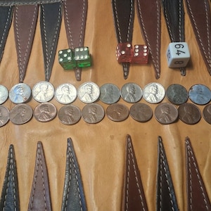 May include: A backgammon board with brown and black triangles, dice, and rows of pennies. Three dice are visible: green, red, and white. The white die shows the number 64. The pennies are arranged in two rows.