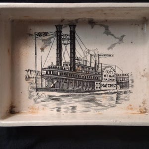 May include: Rectangular ceramic dish with a black and white illustration of the steamboat Natchez. The image includes the name "Natchez" on the flags and the side of the boat. The dish has a cream-colored background with some discoloration.