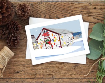May include: A white Christmas card with a red and white illustration of a small, decorated, wooden building with a red lobster on the side. The building is covered in colorful buoys and has a red wreath on the door. The building is set against a blue sky with white clouds and a snowy ground.