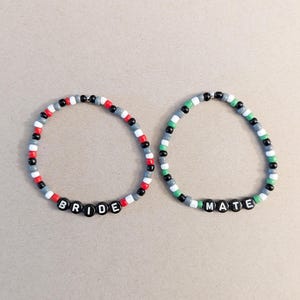 May include: Two beaded bracelets. One bracelet spells "BRIDE" with black letter beads, and the other spells "MATE". Both bracelets have a pattern of red, white, black, and grey beads. The second bracelet has green beads.