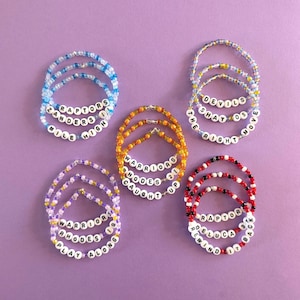 May include: Five sets of circular beaded bracelets featuring white letter beads with black text. The bracelets include phrases such as RAPTORS ZANDERS MILE HIGH, DEVILS SHAY 05 THE RIGHT MOVE, WARRIORS RHODES 21 CAUGHT UP, WARRIORS RHODES 19 PLAY ALONG, and RAPTOR DELUCA 93 REWIND IT BACK.